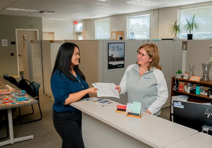 A CSUMB HR representative smiles while handing an employee paperwork across a front desk in a bright and welcoming office, decorated with plants and inspiration posters.