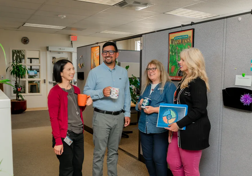 Four CSUMB HR staff members enjoy a casual coffee break together in a collaborative office space, laughing and chatting with colorful mugs and team materials in hand.
