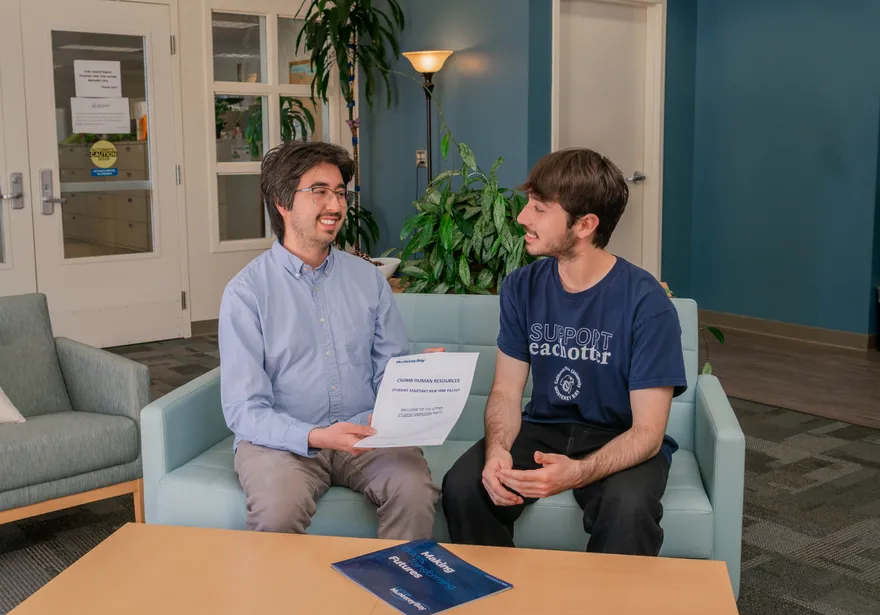 An HR employee and a CSUMB student sit on a couch in the HR reception area reviewing a new hire packet for student assistants, surrounded by welcoming decor and indoor plants.