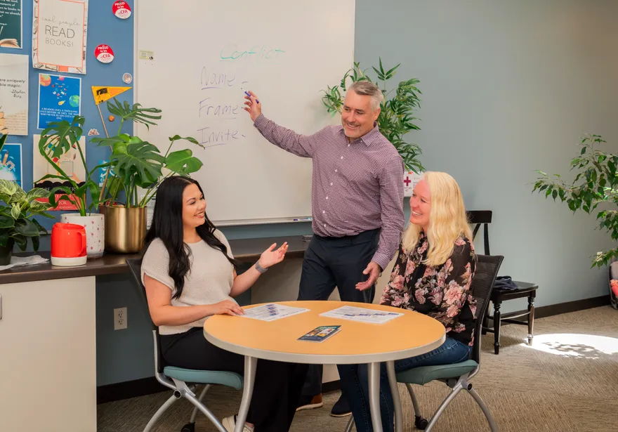 CSUMB Ombuds standing beside a small round table and gesturing to a whiteboard while meeting with two faculty members in an office.
