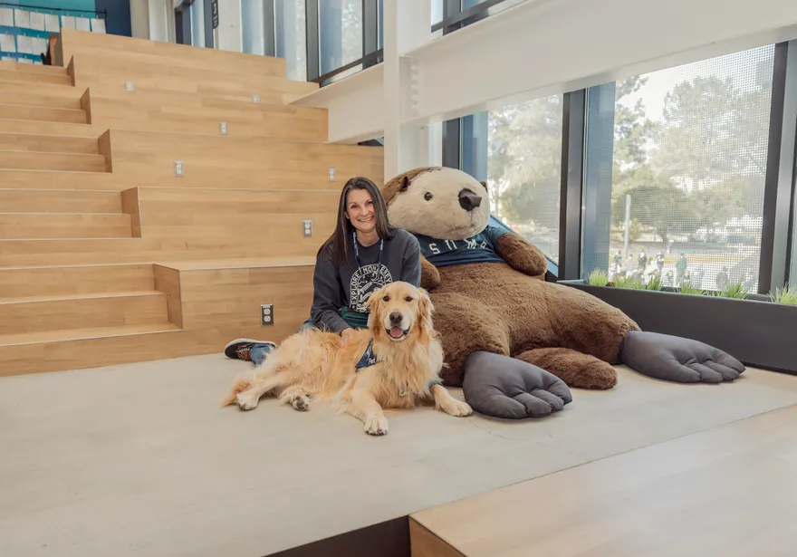 Blue the therapy laying down in front of the large Monte plushe in the OSU with his owner sitting behind him.