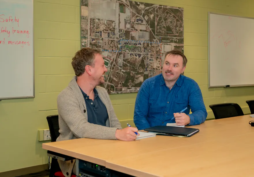 Two staff members talk while seated at a conference table with a campus map on the wall. One is mid-conversation while the other smiles and listens.