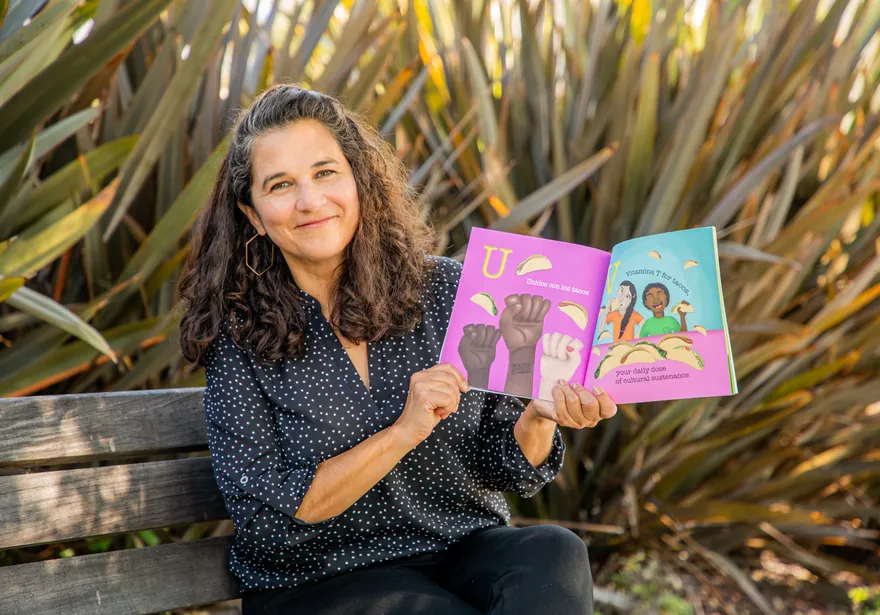A woman sitting on a bench outdoors smiles while holding open a colorful children’s book featuring illustrations of raised fists and people eating tacos, with text in both English and Spanish.