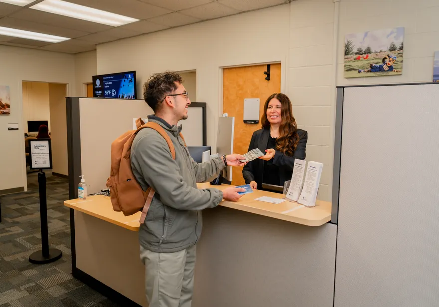 Inside the Bursar’s Office at �鶹��, a student makes a cash payment to a smiling finance staff member across the front counter.