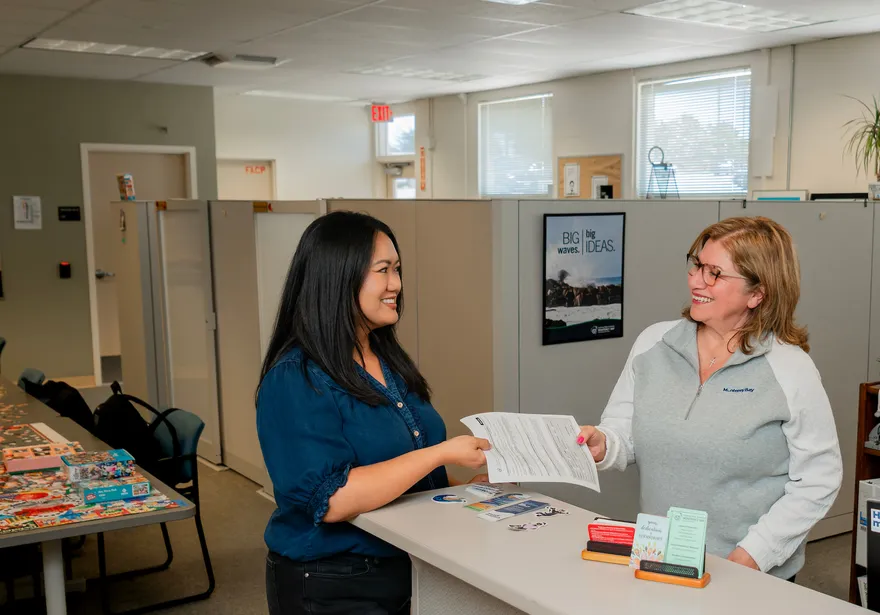 A ���ӽ紫ý HR representative smiles while handing an employee paperwork across a front desk in a bright and welcoming office, decorated with plants and inspiration posters.
