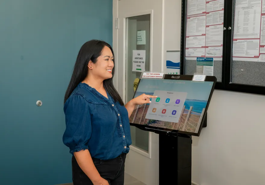 An employee interacts with a digital HR kiosk at ���ӽ紫ý, smiling while selecting options on the touchscreen labeled “Welcome to ���ӽ紫ý HR.”