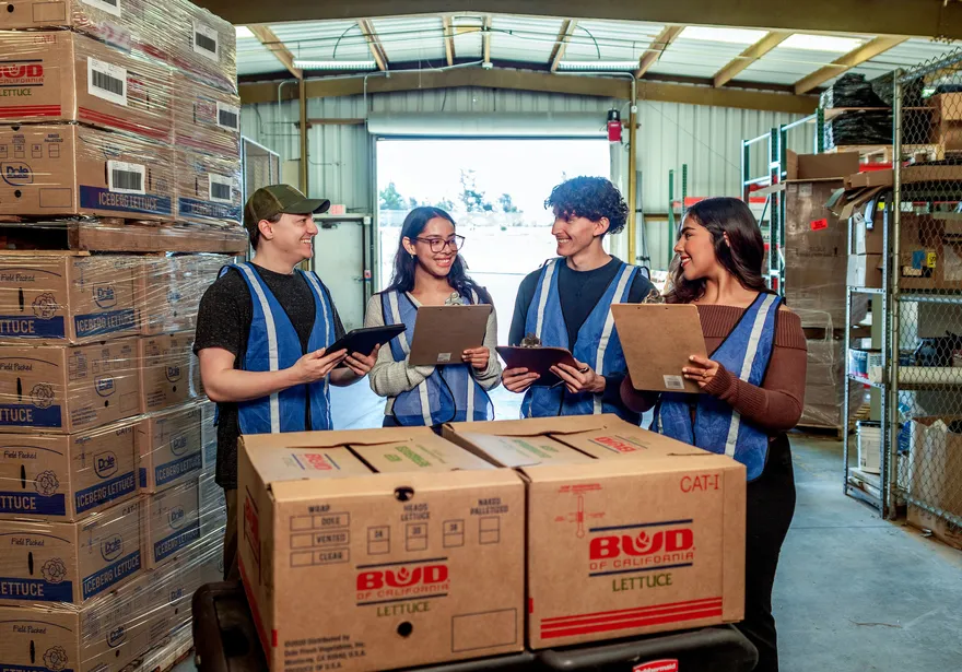 Group of students standing next to each other talking in a warehouse holding clipboards.
