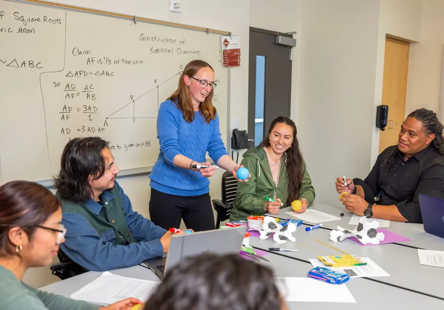 Professor standing near the students sitting at a table looking at her.