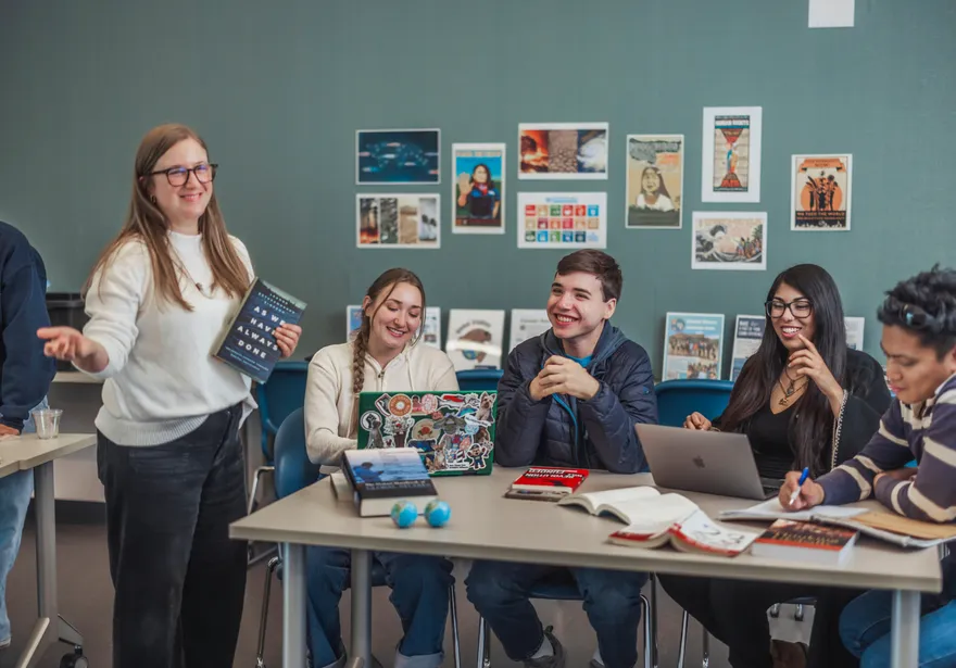 A group of four students sitting at a table doing work and smiling while the professor stands near them smiling and holding a book