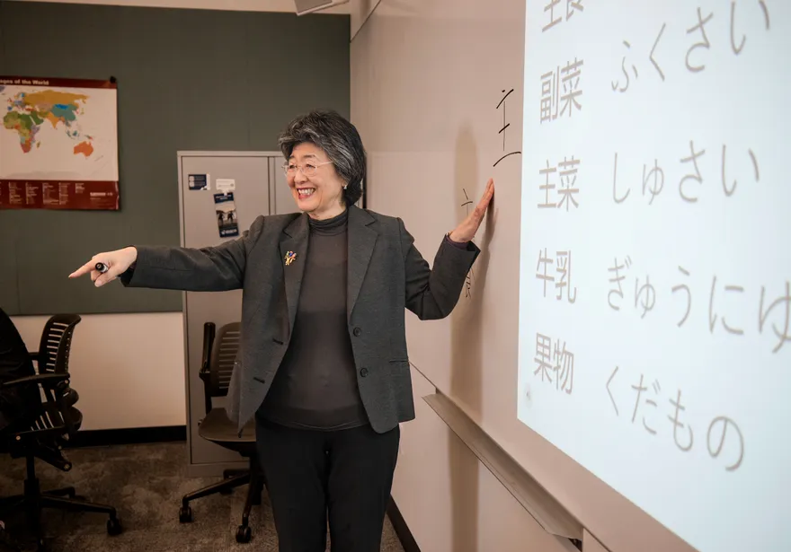 Professor standing in front of a white board pointing at Japanese symbols and smiling while teaching the class