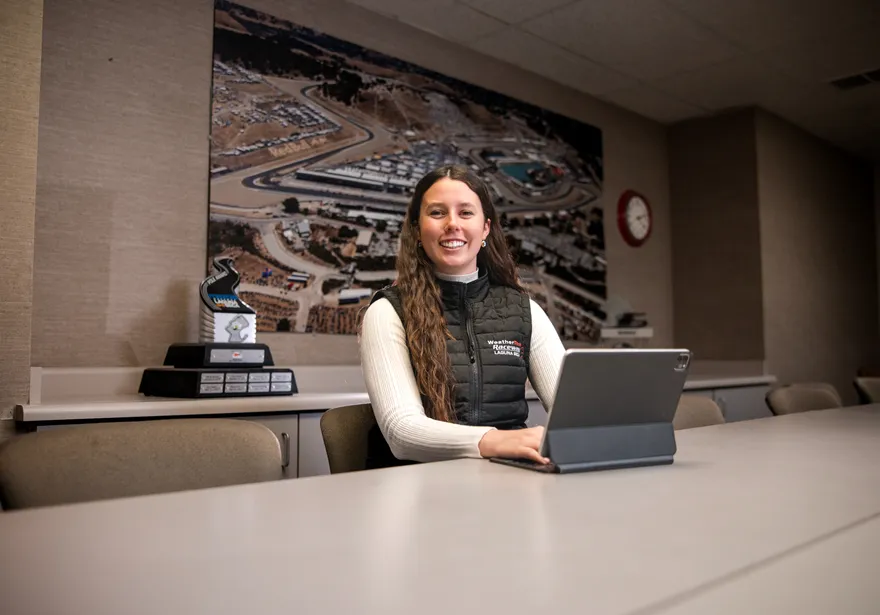 A student sitting at a desk smiling at the camera while she has her computer open on the desk.