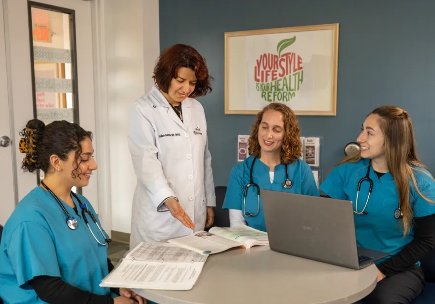 A group of three nursing students in scrubs sitting down at a table listening to a professor