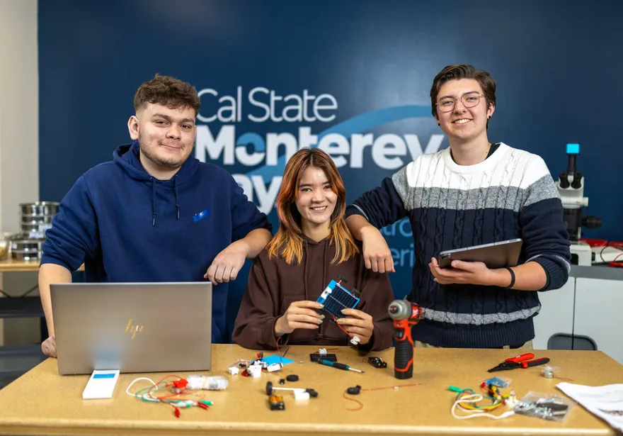 A group of three mechatronics students behind a mechatronics display table smiling at the camera