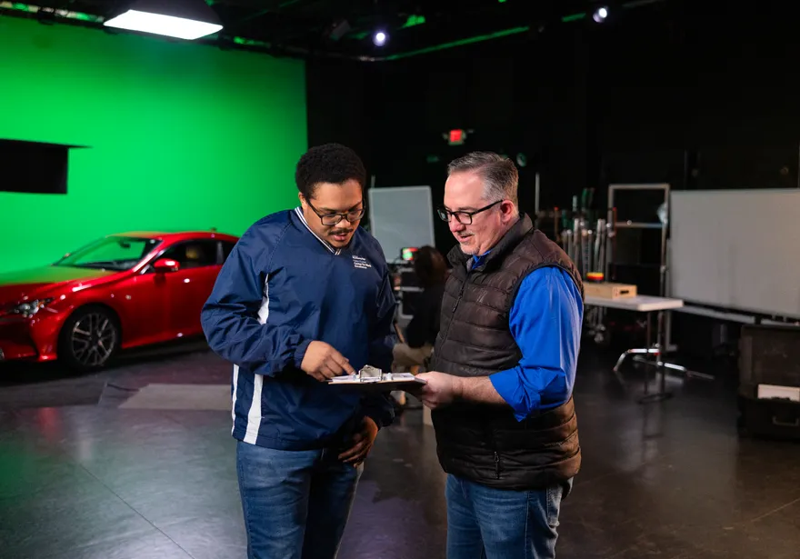 Two film crew members holding a clipboard and discussing in front of a green screen and a red car behind them.