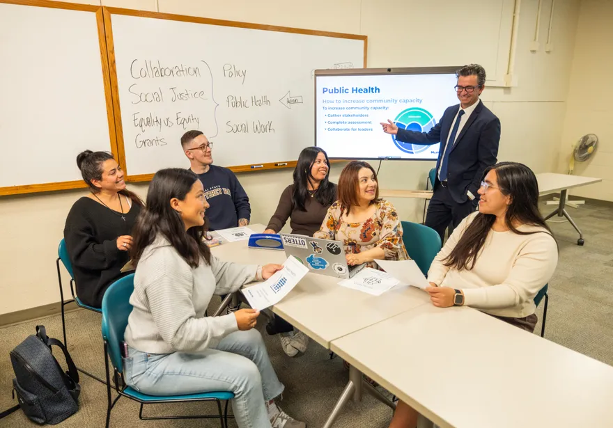 Six students sitting at a table listening and taking notes while a professor stands, teaching them in front of a white board and screen.