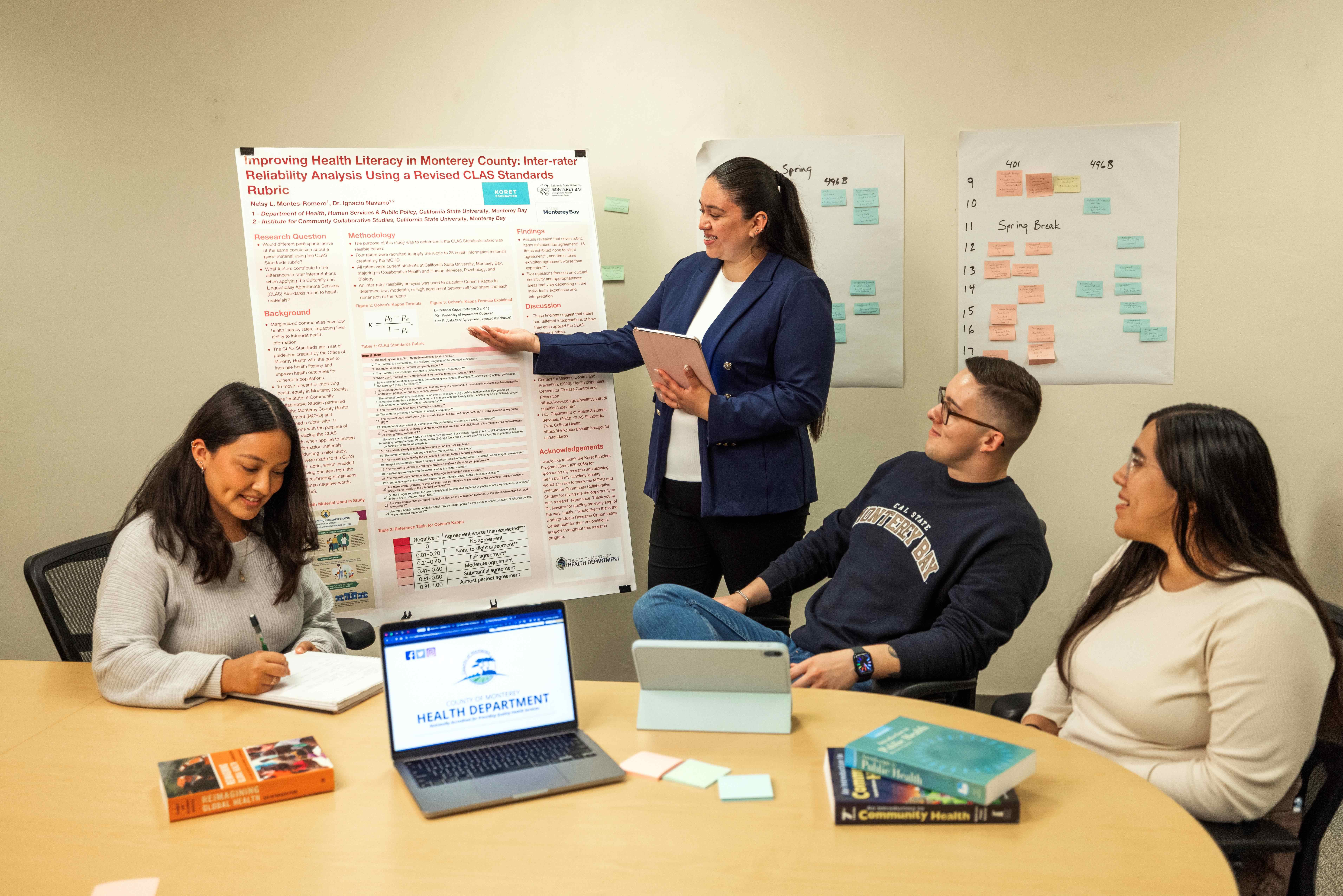 A student presenting a poster of work to three other students who are sitting at a table listening and taking notes.